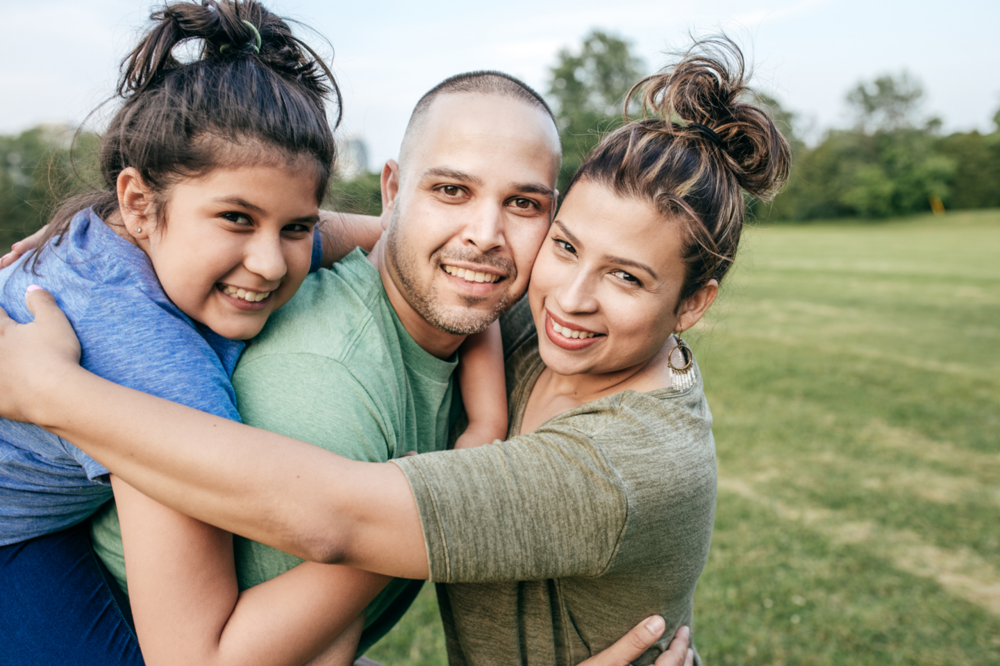 Familia de tres personas, sonriente y abrazada, en un hermoso campo