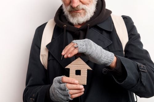 Older man with beard wearing fingerless gloves holds cardboard picture of house