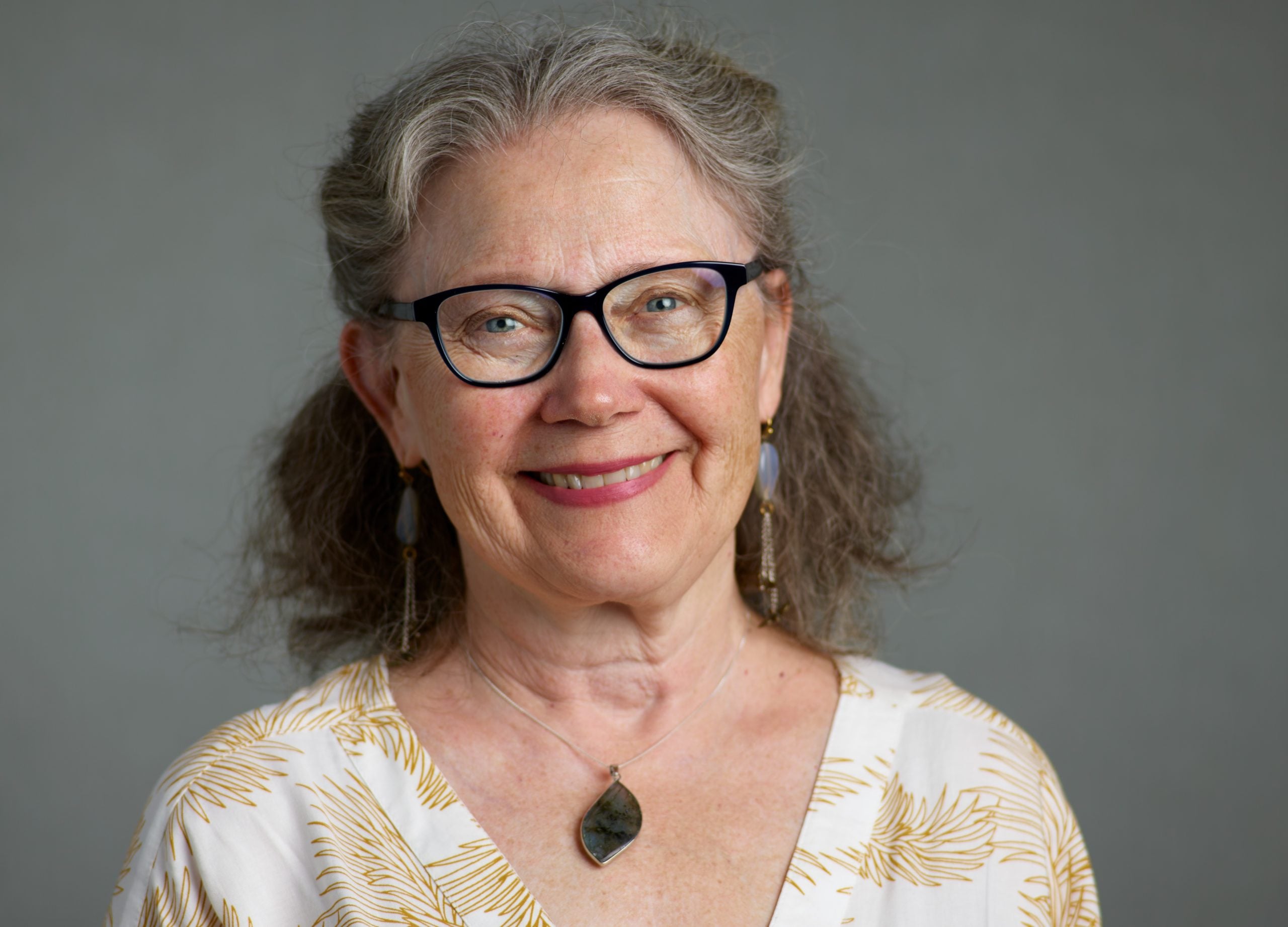 Formal portrait of Bee, smiling with leaf-shaped necklace before a gray background