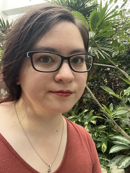 Luci in black framed glasses looks into camera in front of a large stand of leafy plants