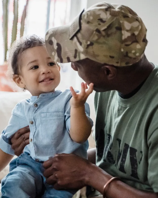 Man in camouflage cap faces toward smiling child touching his face