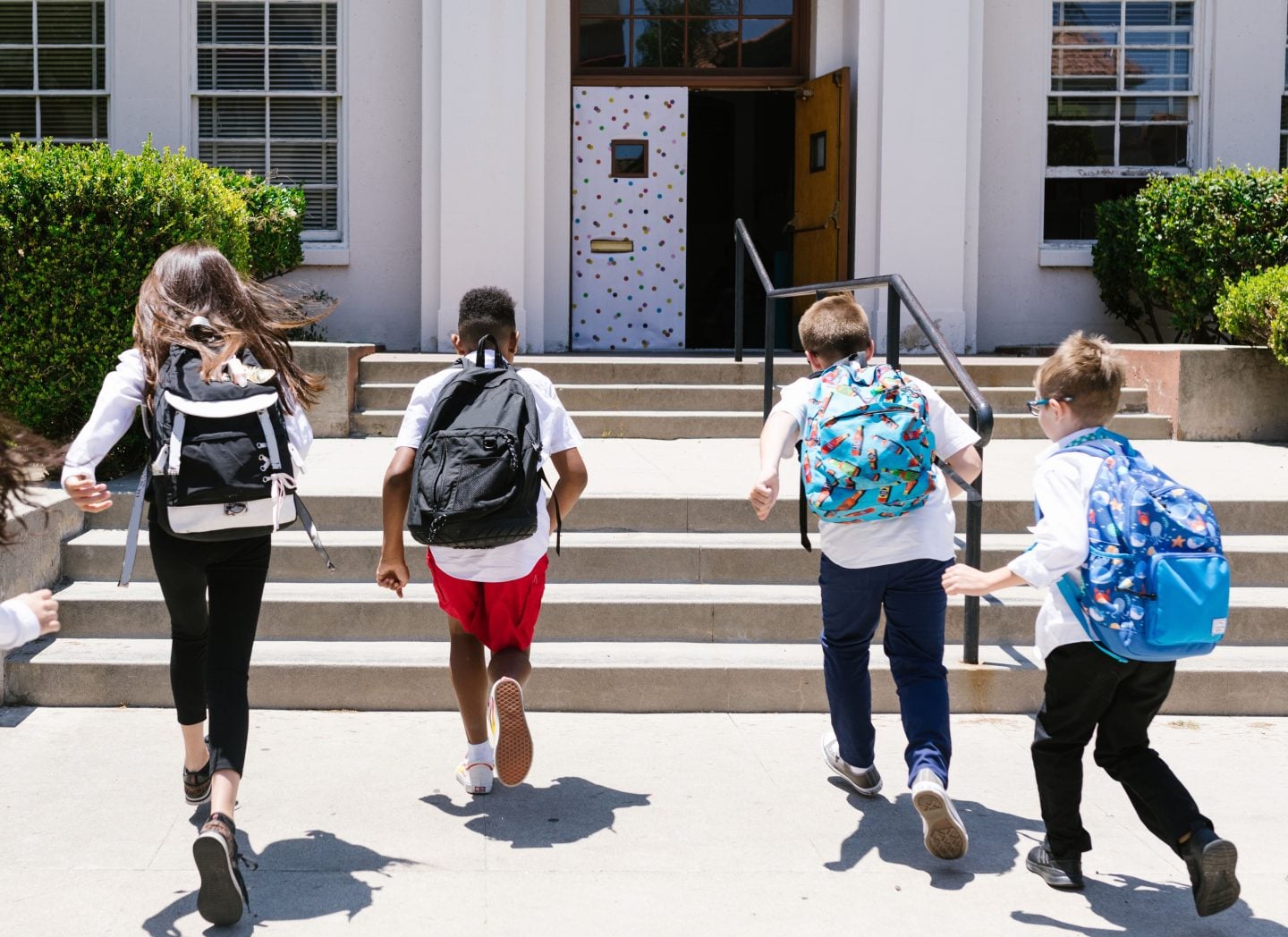 Four students in backpacks run up the steps to an open school door